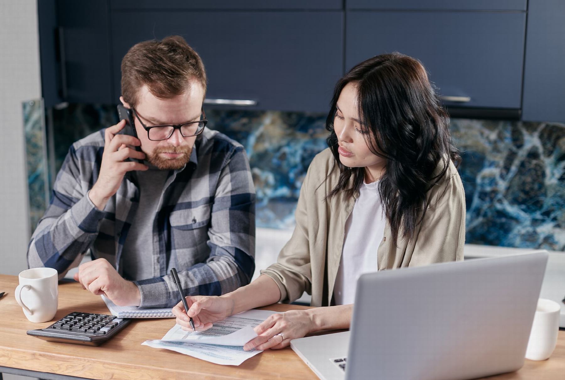 Un homme au téléphone et une femme qui annote un document, à une table de cuisine, avec un ordinateur portable et une calculatrice.