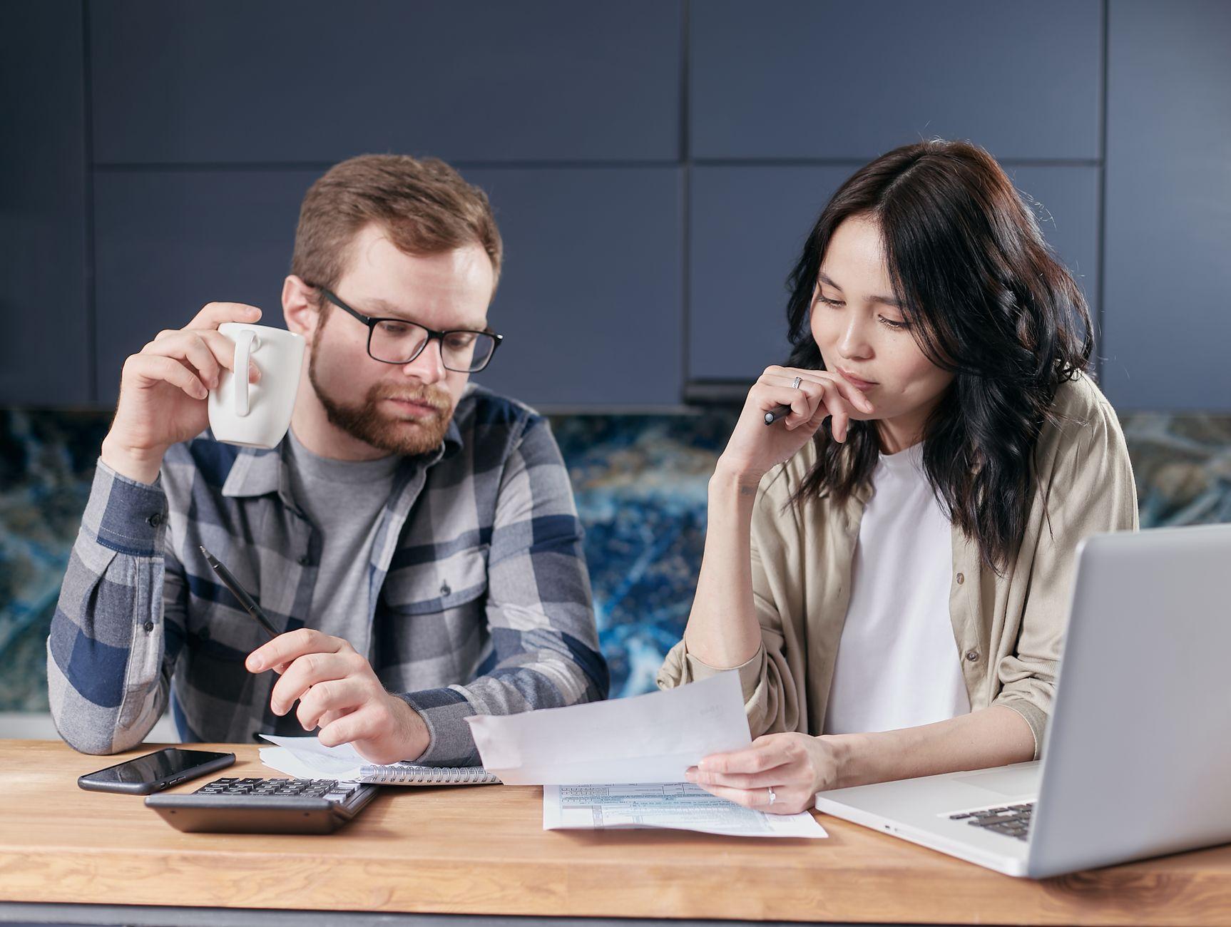 Un couple attablé dans une cuisine, une tasse à la main, une feuille de facture sous les yeux, à côté d’un ordinateur portable et d’une calculatrice.