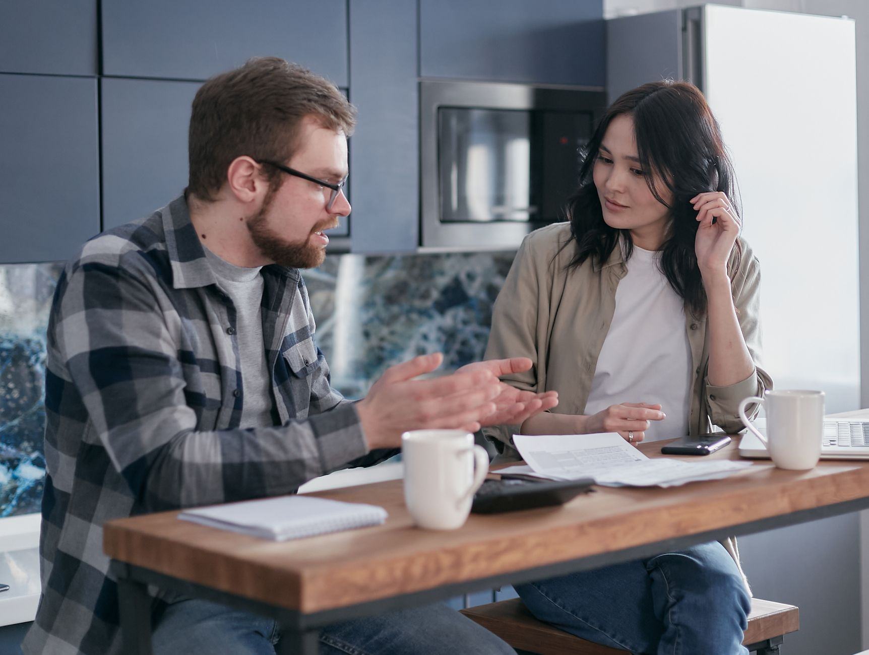 Un homme et une femme discutent à une table de cuisine, des documents, une calculatrice et des tasses devant eux.