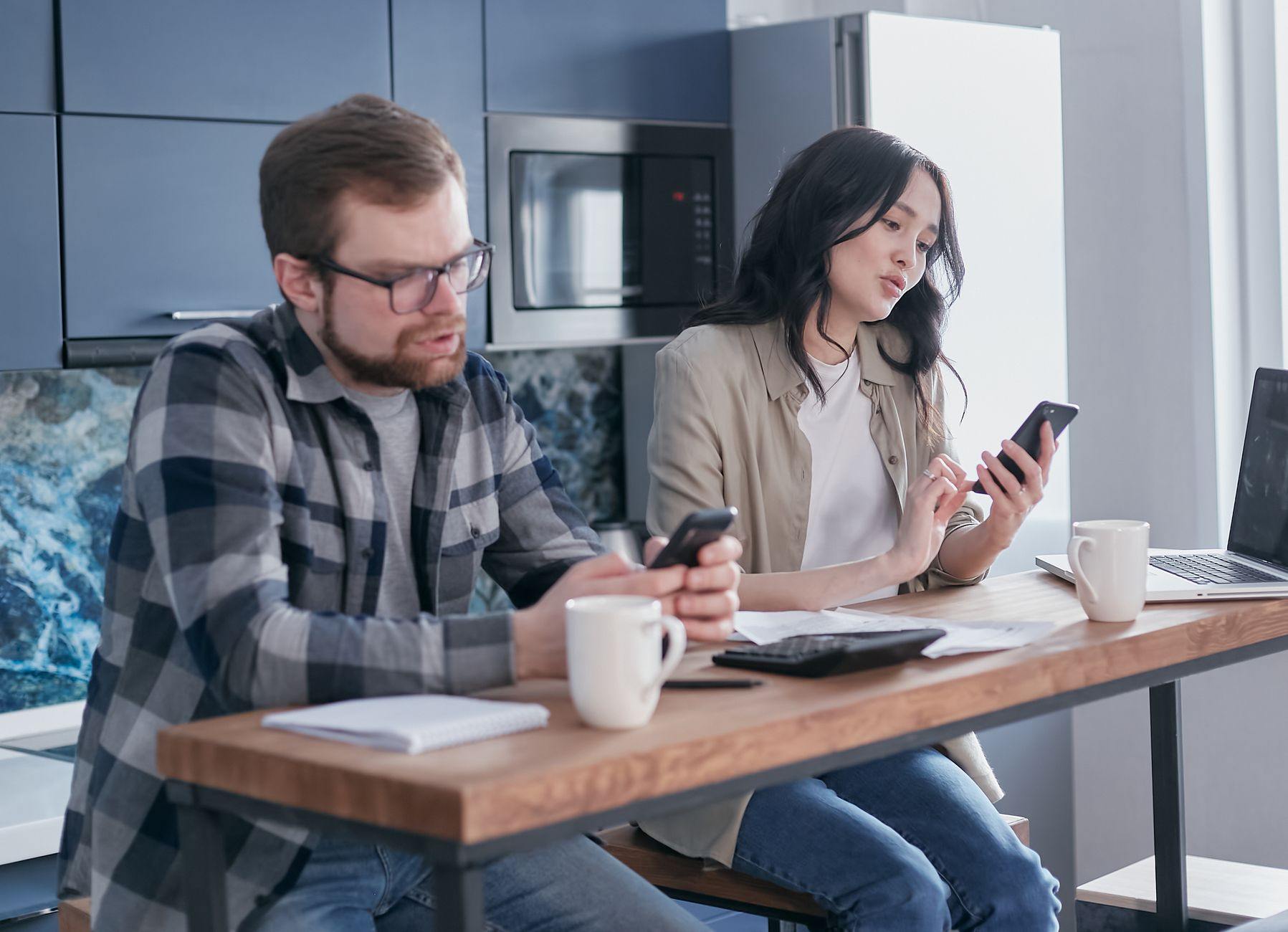Un homme et une femme assis à une table de cuisine, chacun un smartphone en main, avec des documents et un ordinateur portable à côté.
