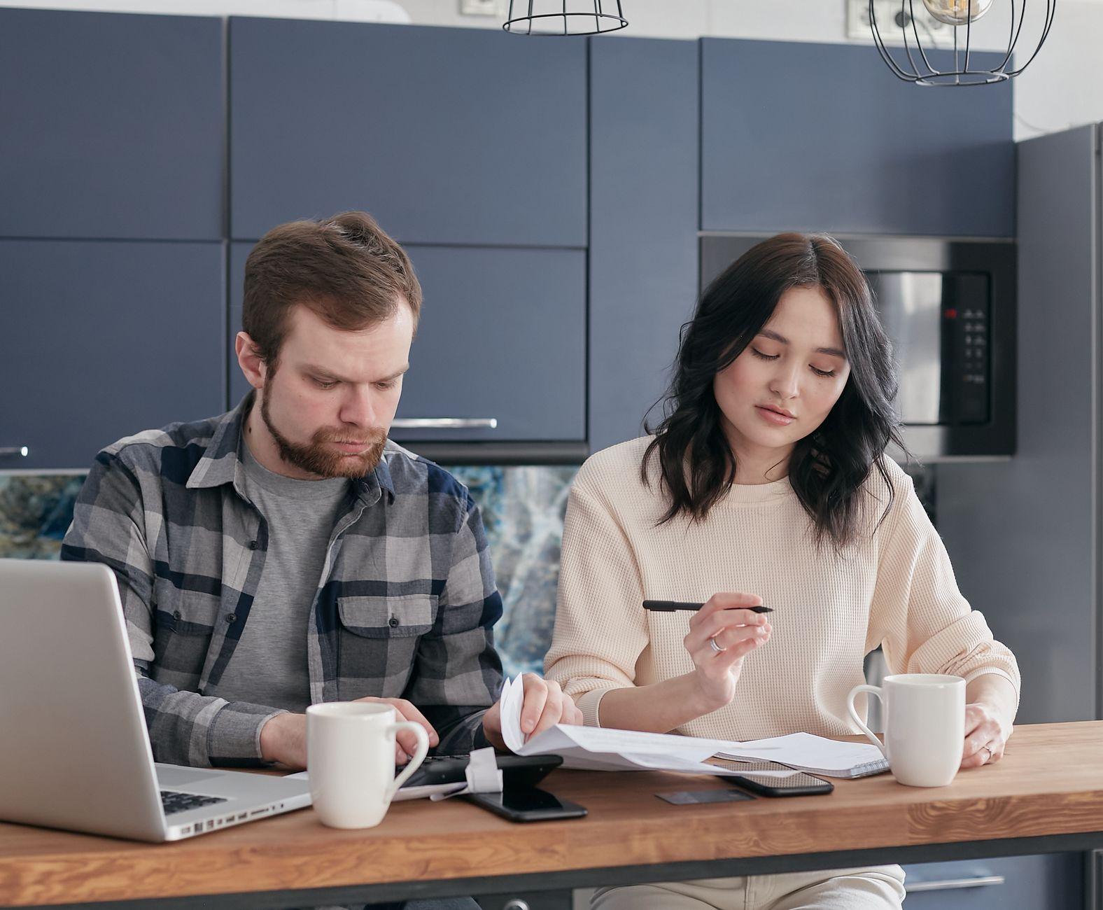 Un homme et une femme à une table de cuisine, un ordinateur portable ouvert et des documents devant eux.